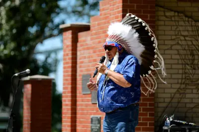 alt="Elder Charlie Fox wears a large feather headdress while speaking into a microphone"